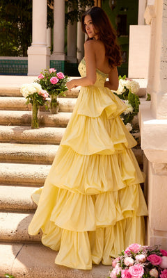 A woman in the Ava Presley 26417 long prom dress—a strapless, tiered yellow ruffle gown—stands on stone steps, holding a floral bouquet with more flower arrangements beside her.