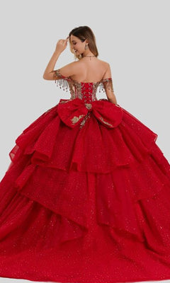 A woman stands against a plain gray background, shown from the back, wearing the Quinceanera Dress 26417 by Stella Couture—an off-shoulder, layered red ball gown adorned with a large bow and gold detailing, featuring a gold-embellished corset bodice.