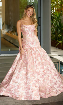 A woman poses indoors in the Ava Presley 26419 Drop-Waist Long Print Prom Gown in blush, featuring thin straps and a floor-length skirt, smiling near glass doors and greenery.