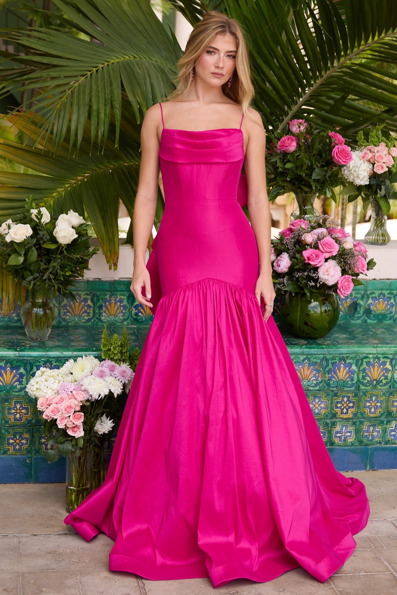 A woman poses before lush greenery and blooms, wearing the Ava Presley 26422 drop-waist mermaid prom gown in bright pink, featuring a fitted bodice and flared skirt.