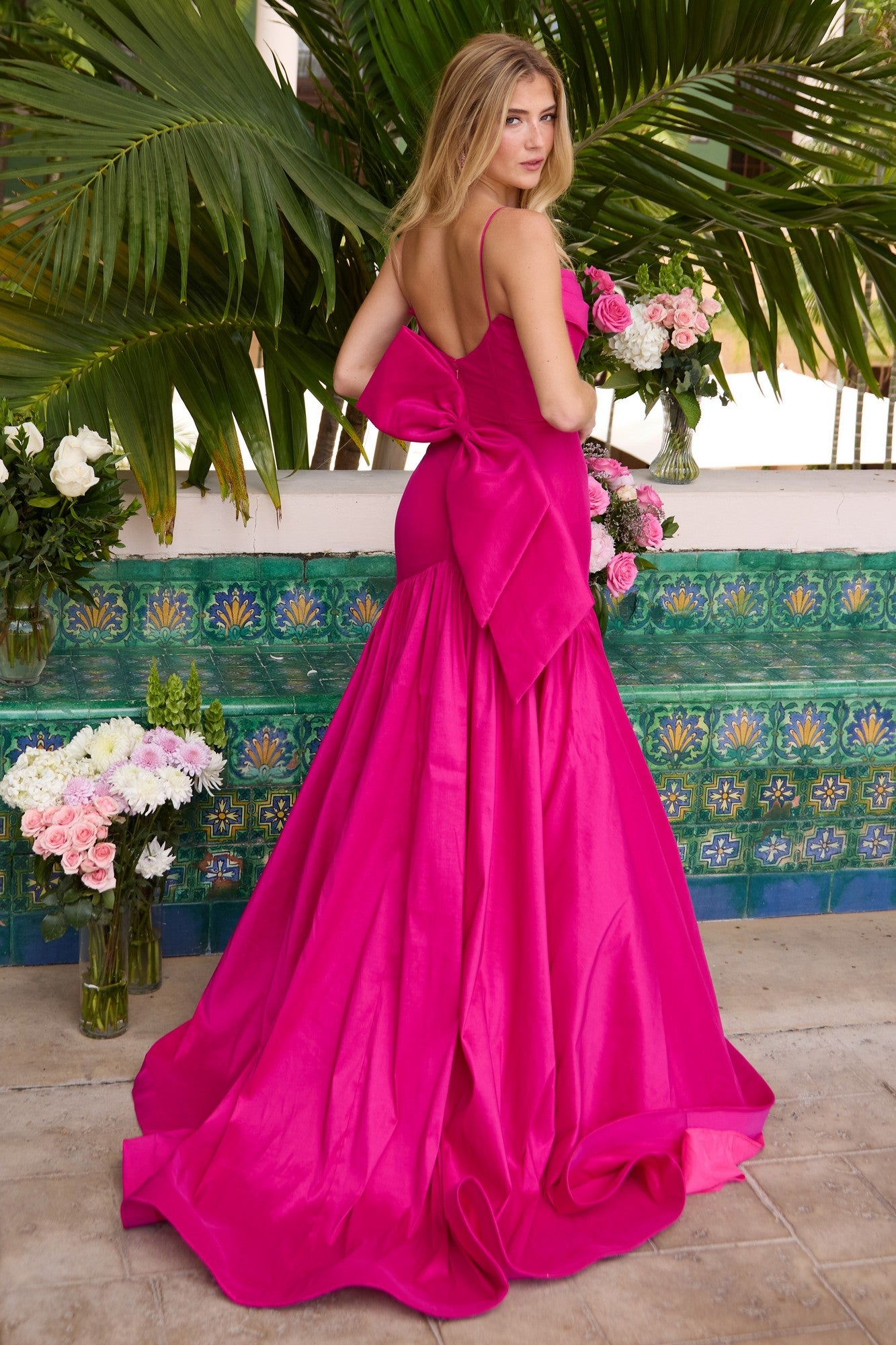 A woman in the bright pink Drop-Waist Mermaid Prom Gown by Ava Presley 26422 stands before green tiles and palm leaves, holding a bouquet of flowers.