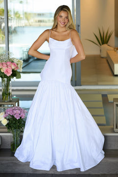 A woman stands indoors smiling with hands on hips, wearing the Ava Presley 26422 Drop-Waist Mermaid Prom Gown. Flowers and modern decor accent the background.