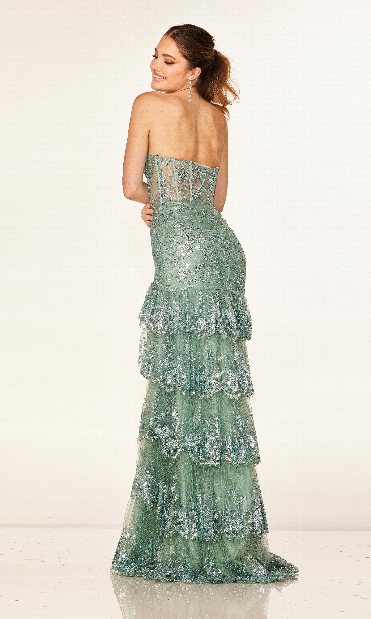 A woman models the Cecilia Couture 2642, a strapless green sequined long prom dress with a corset bodice and tiered layered skirt, standing against a plain light background and smiling over her shoulder.