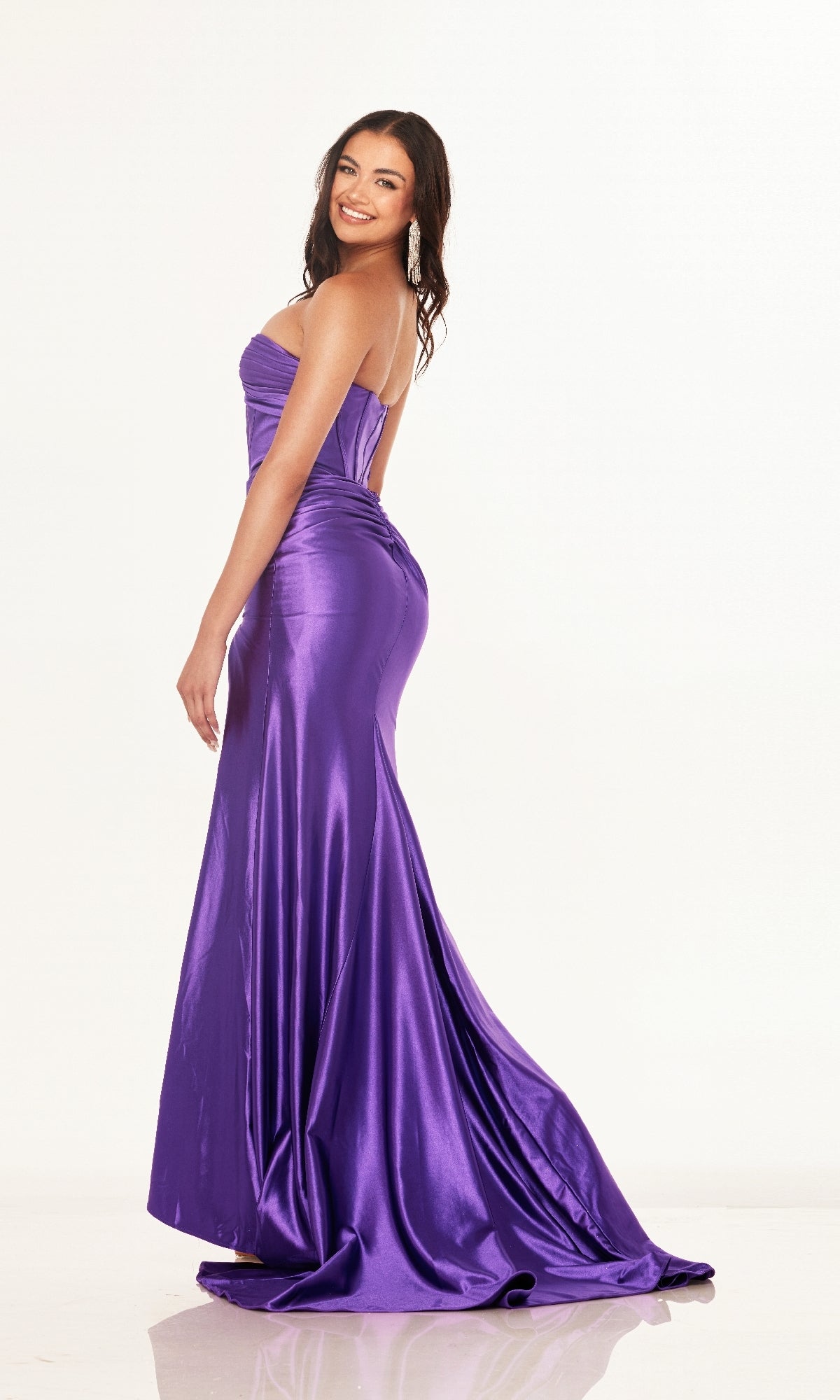 A woman smiles sideways in the Cut-Out Strapless Prom Gown: Cecilia Couture 2643, featuring a corset-style bodice and shiny purple, floor-length skirt, posing against a plain white background.