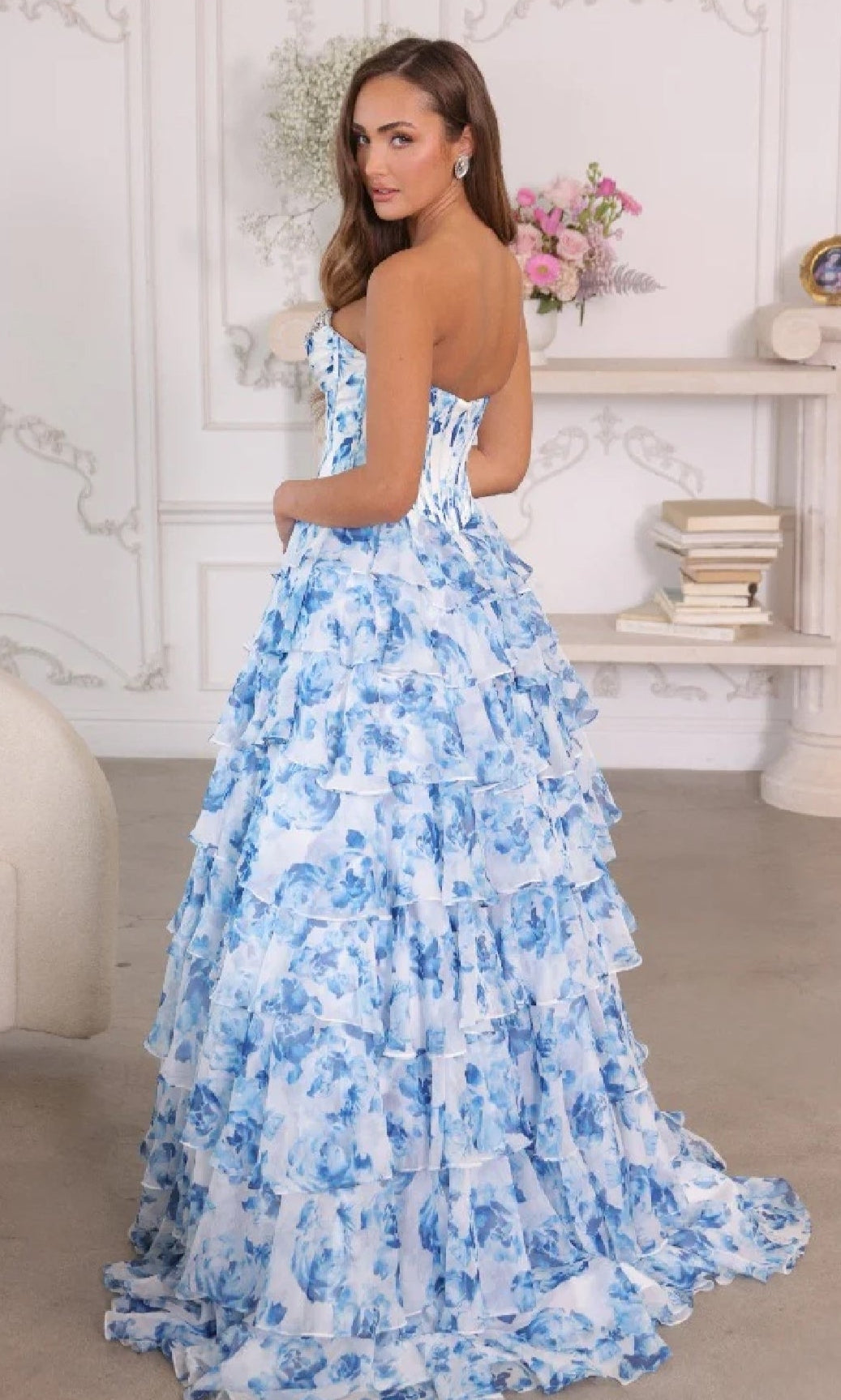A woman wearing the Ava Presley 26441 long prom dress with a strapless bodice and blue and white floral ruffle ball-gown skirt stands indoors, glancing over her shoulder with shelves of books and a vase of flowers in the background.