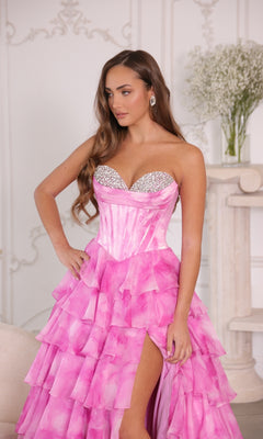 A woman stands indoors with one hand on her hip, wearing the Ava Presley 26442 long prom dress—a strapless pink ball gown with a tiered ruffle skirt. A vase of white flowers sits on a shelf behind her.