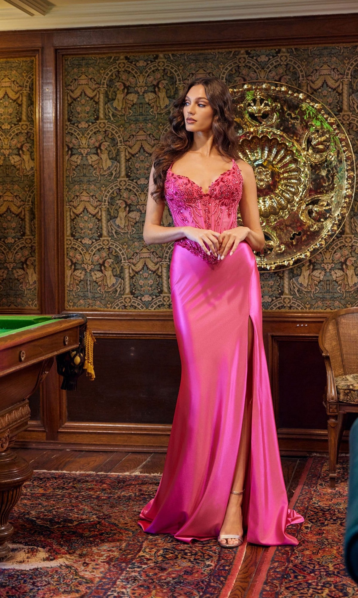 A woman wears the Juliet JT2645K long prom dress, a fitted, floor-length pink gown with a thigh-high slit, standing in an ornate room with wood paneling and decorative tapestry.