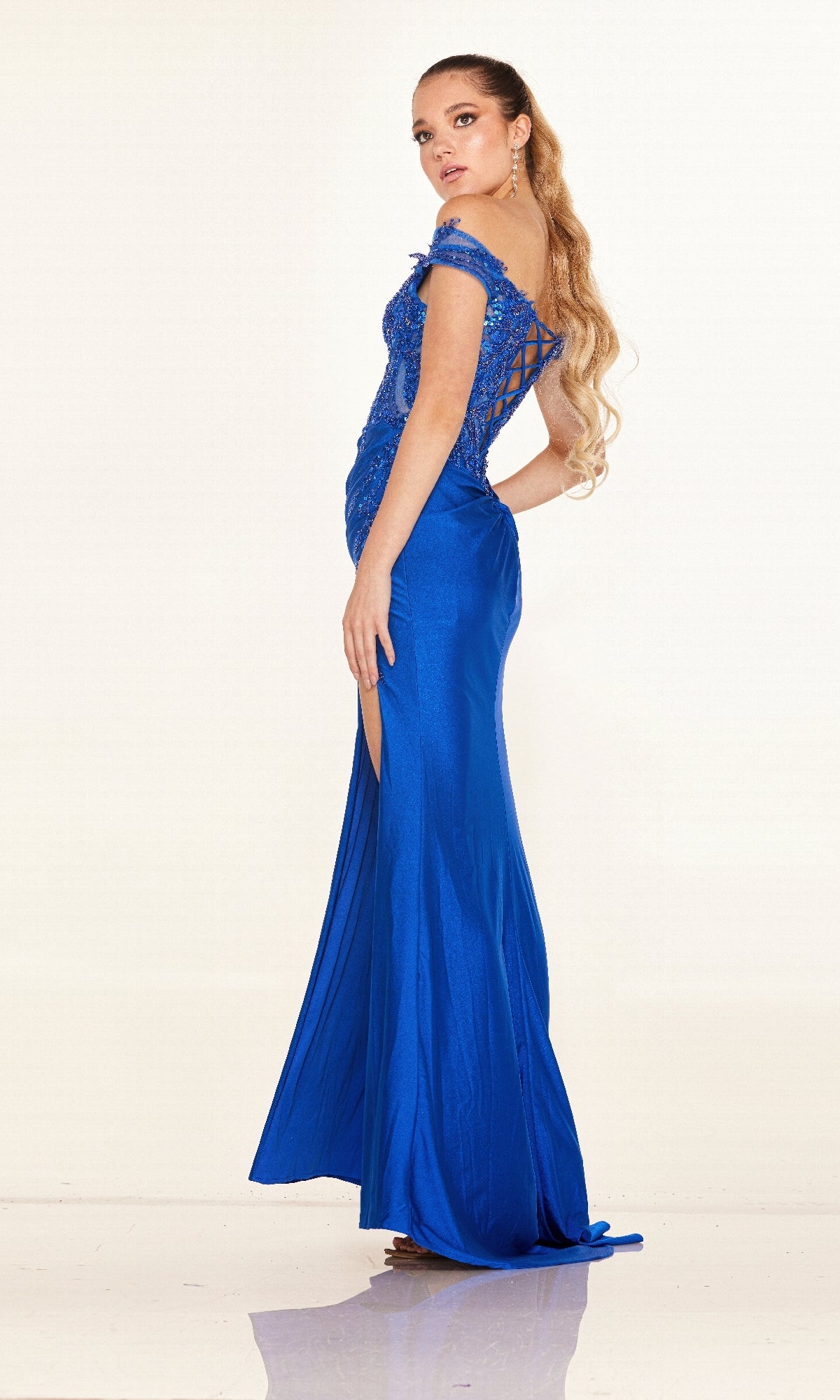 A woman stands sideways against a plain, light background wearing the Cecilia Couture 2645 long prom dress in blue, featuring off-the-shoulder lace details and a delicate lace-up back.