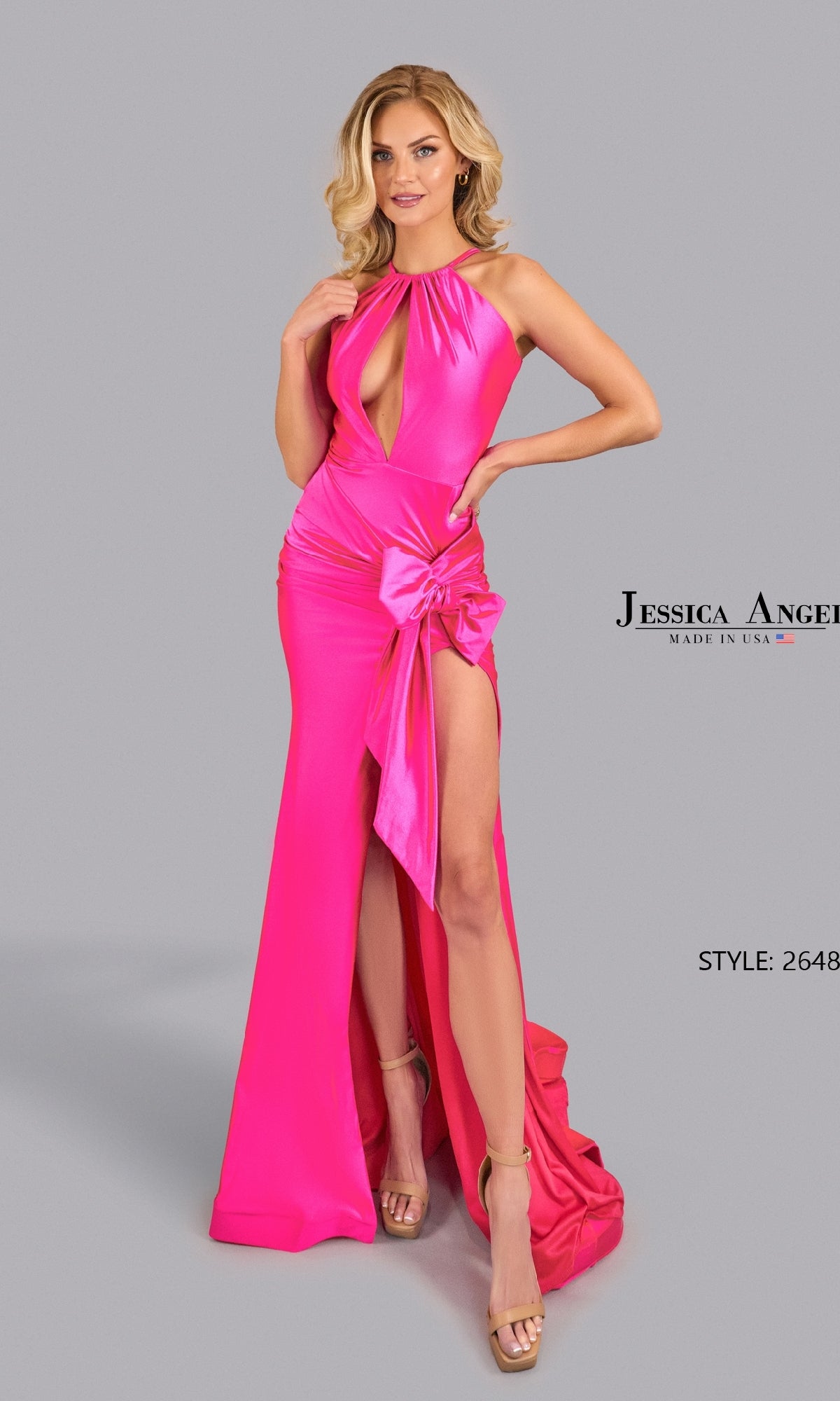 A woman in a bright pink Cut-Out Sexy Long Prom Dress: Jessica Angel 2648, featuring a front cutout, high slit, and bow detail, stands against a plain gray background.