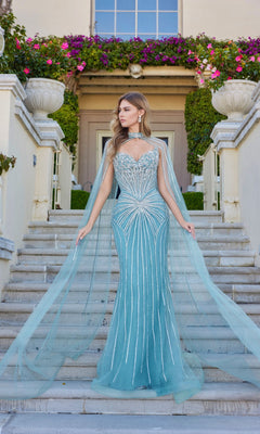 A woman in the Juliet JT2649K long prom dress in deep sage, featuring a strapless design and sheer floor-length chiffon cape with hot-stone accents, stands outdoors on stairs surrounded by flowers and greenery.