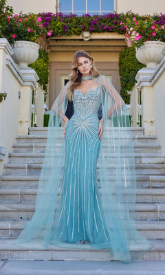 A woman stands on stone steps in the Juliet JT2649K Long Prom Dress, a fitted light blue gown with hot-stone embellishments and sheer cape sleeves, with a building and flowers in the background.