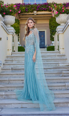 A woman in the Juliet JT2649K long prom dress—light blue, strapless, with intricate beading and a sheer, floor-length chiffon cape—stands on outdoor stone steps before a building decorated with flowers and greenery.
