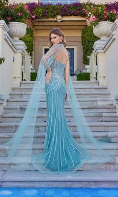 A woman wearing the Juliet JT2649K long prom dress—an elegant teal strapless gown with silver accents and a floor-length chiffon cape—stands on outdoor steps before a flower-adorned building.