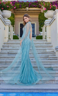 A woman in the Juliet JT2649K long prom dress, featuring a fitted light blue design with silver accents and a floor-length chiffon cape, stands on stone steps outside a flower-adorned building.