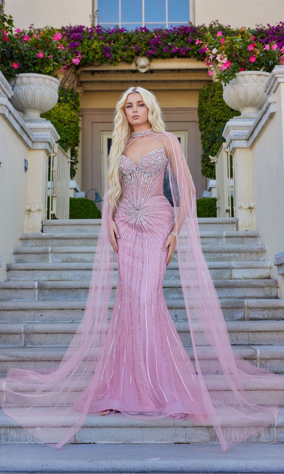 Wearing the Juliet JT2649K long prom dress in rose, a woman stands on outdoor stone steps before a flower-adorned building; the fitted, embellished strapless gown features a sheer, floor-length chiffon cape.