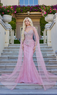 Wearing the Juliet JT2649K long prom dress in rose, a woman stands on outdoor stone steps before a flower-adorned building; the fitted, embellished strapless gown features a sheer, floor-length chiffon cape.