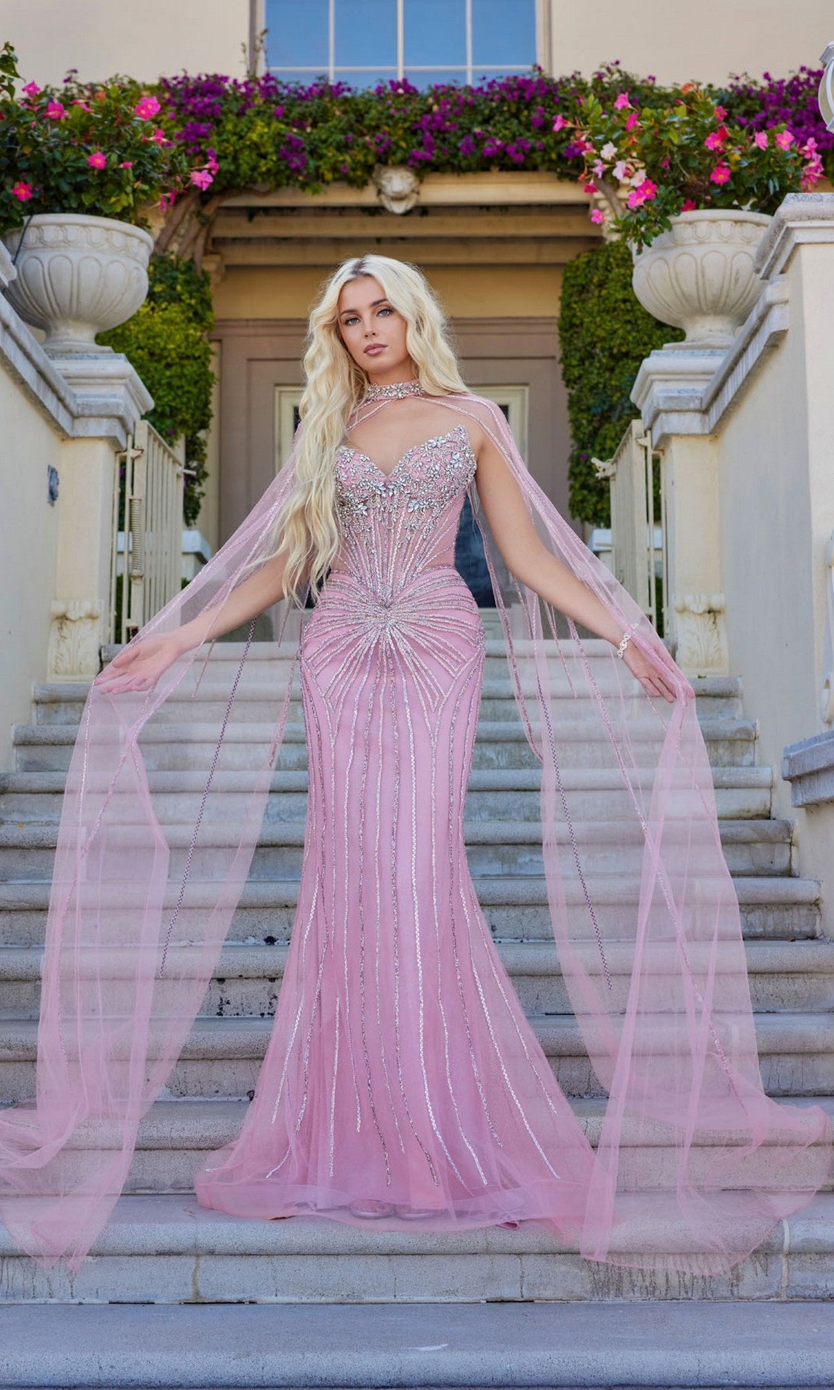A person in the Juliet JT2649K long prom dress—a fitted, light pink gown with hot-stone details and a sheer, floor-length chiffon cape—stands on stone steps before a flower-adorned building.