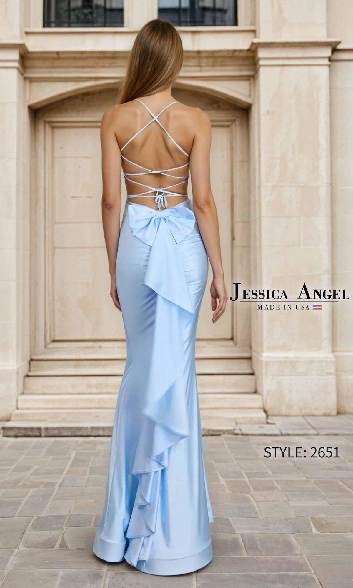 A woman stands outdoors wearing the Jessica Angel 2651 long prom dress, a light blue, floor-length, sleeveless gown featuring crisscross lace-up back straps and a large bow.