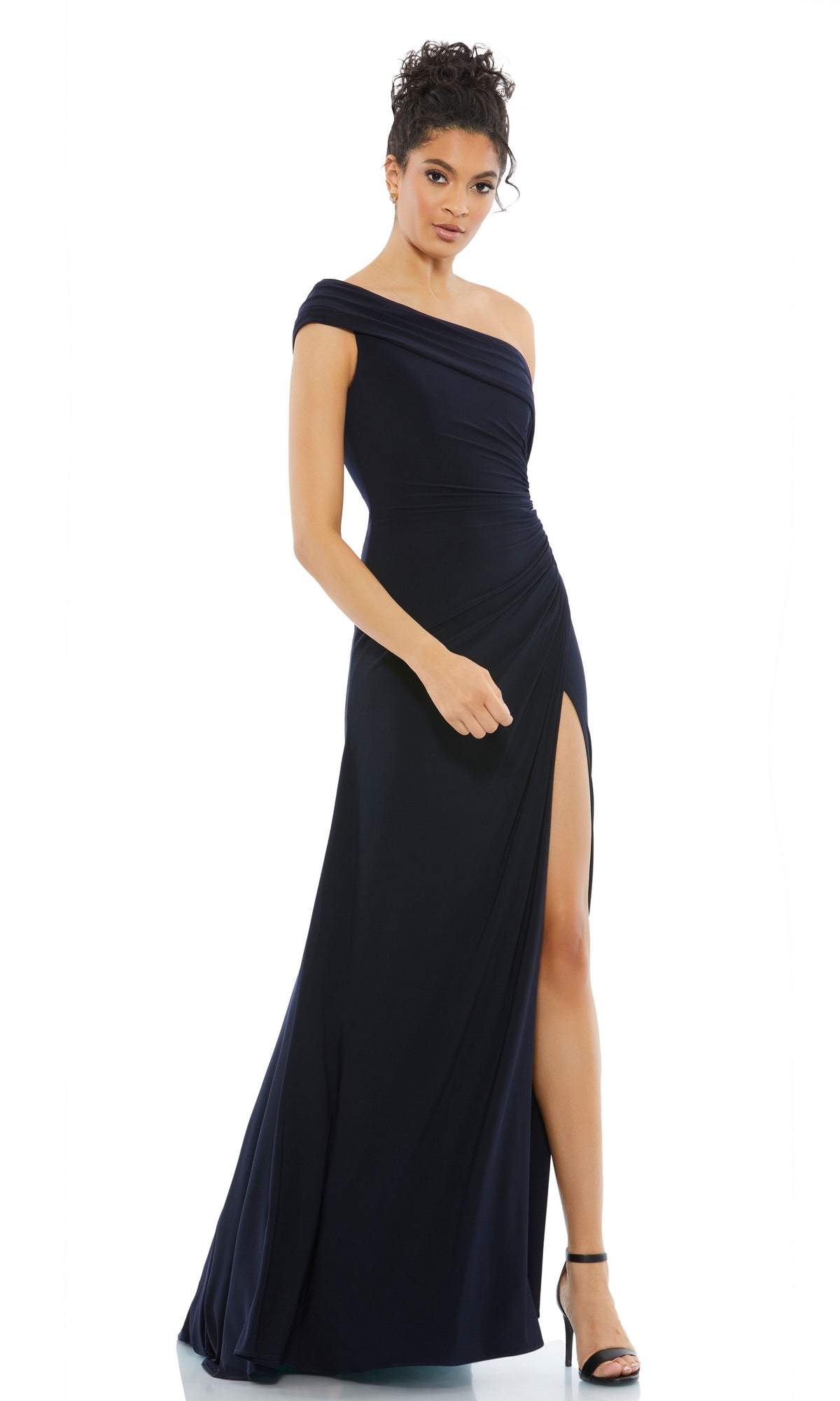 A woman in the Mac Duggal 26517 One-Shoulder Long Prom Dress, featuring a thigh-high slit and navy hue, pairs the gown with black high-heeled sandals while standing against a white background.