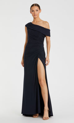A woman stands against a plain light background wearing the Mac Duggal 26517 One-Shoulder Long Prom Dress in navy, featuring a high thigh slit and nude heeled sandals.