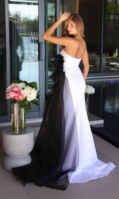 A woman in the Ava Presley 26518 long prom dress, featuring a strapless design with a large black organza bow and train, stands by a glass door near floral arrangements and a pool.
