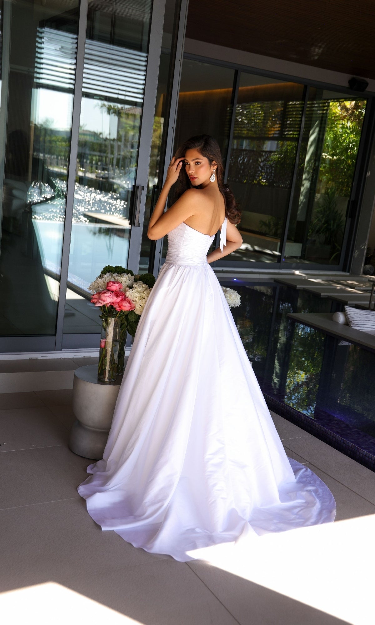 A woman wearing the Ava Presley 26524 Long Prom Dress stands indoors by a glass door, next to a vase of pink and white flowers, looking over her shoulder.