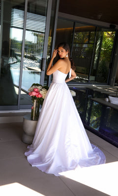 A woman wearing the Ava Presley 26524 Long Prom Dress stands indoors by a glass door, next to a vase of pink and white flowers, looking over her shoulder.