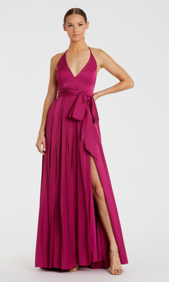 A woman in the Mac Duggal 26531 Bowed Long Halter Formal Dress—sleeveless, magenta, backless, deep v-neckline, waist sash bow, high leg slit—and beige heeled sandals stands against a plain background.