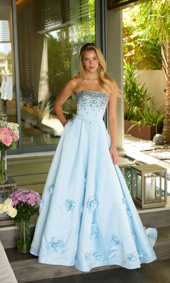 A woman in the Ava Presley 26532 long prom dress, featuring a strapless satin design with 3D floral appliques, stands indoors near large windows and a bouquet of flowers.
