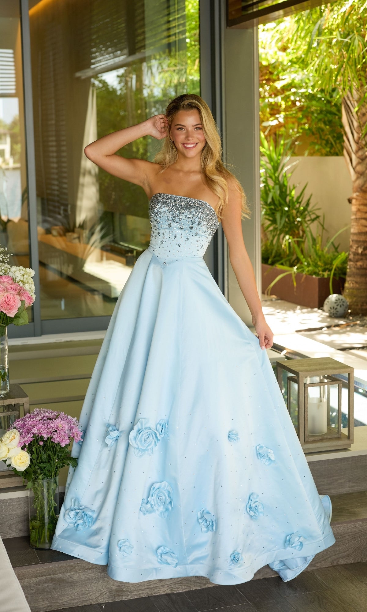 A woman smiles indoors near a window, wearing the Ava Presley 26532 long prom dress with a strapless satin design, beaded bodice, and 3D floral appliques.