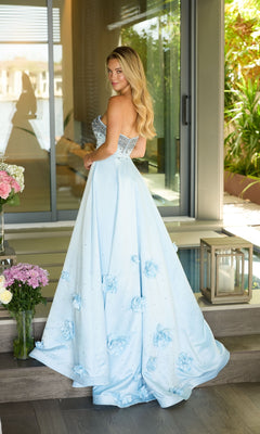 A woman in the Ava Presley 26532 long prom dress, featuring a strapless satin design with a beaded bodice and 3D floral appliques, stands indoors near a glass door amidst flowers and lanterns.