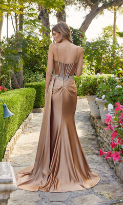 A woman stands on a stone path in a garden, wearing the Juliet JT2653K Long Prom Dress—a copper-colored satin, floor-length gown with thin straps and decorative back detailing.