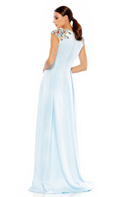 A woman stands with her back mostly to the camera, wearing the Mac Duggal 26540 cap-sleeve long formal dress in light blue, featuring a floor-length A-line silhouette and colorful embroidered shoulder flowers.