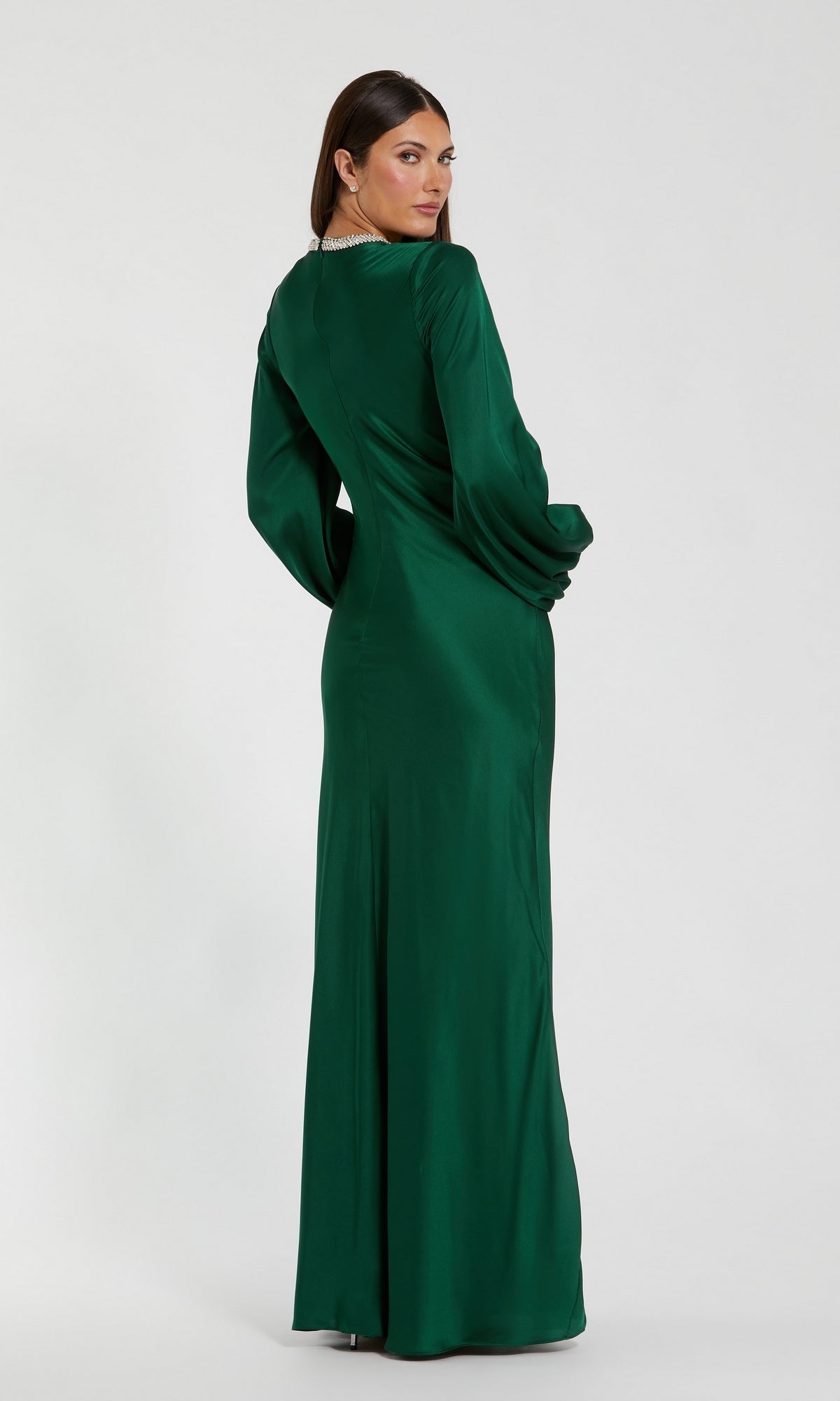Woman wearing the Mac Duggal 26575 Long Sleeve Long Formal Dress in green satin stands facing away, glancing over her shoulder against a plain white background.