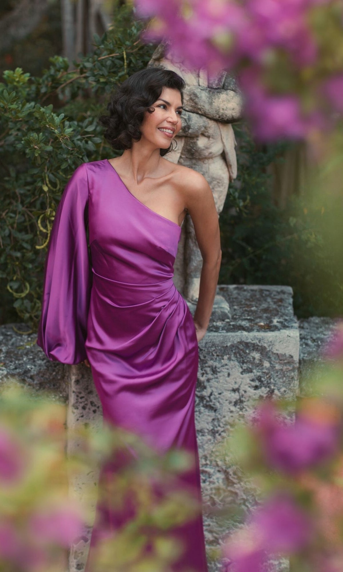 A woman in the Mac Duggal 26588 One-Sleeve Long Formal Dress stands outdoors near a stone structure, surrounded by greenery and purple flowers, smiling and looking to the side.