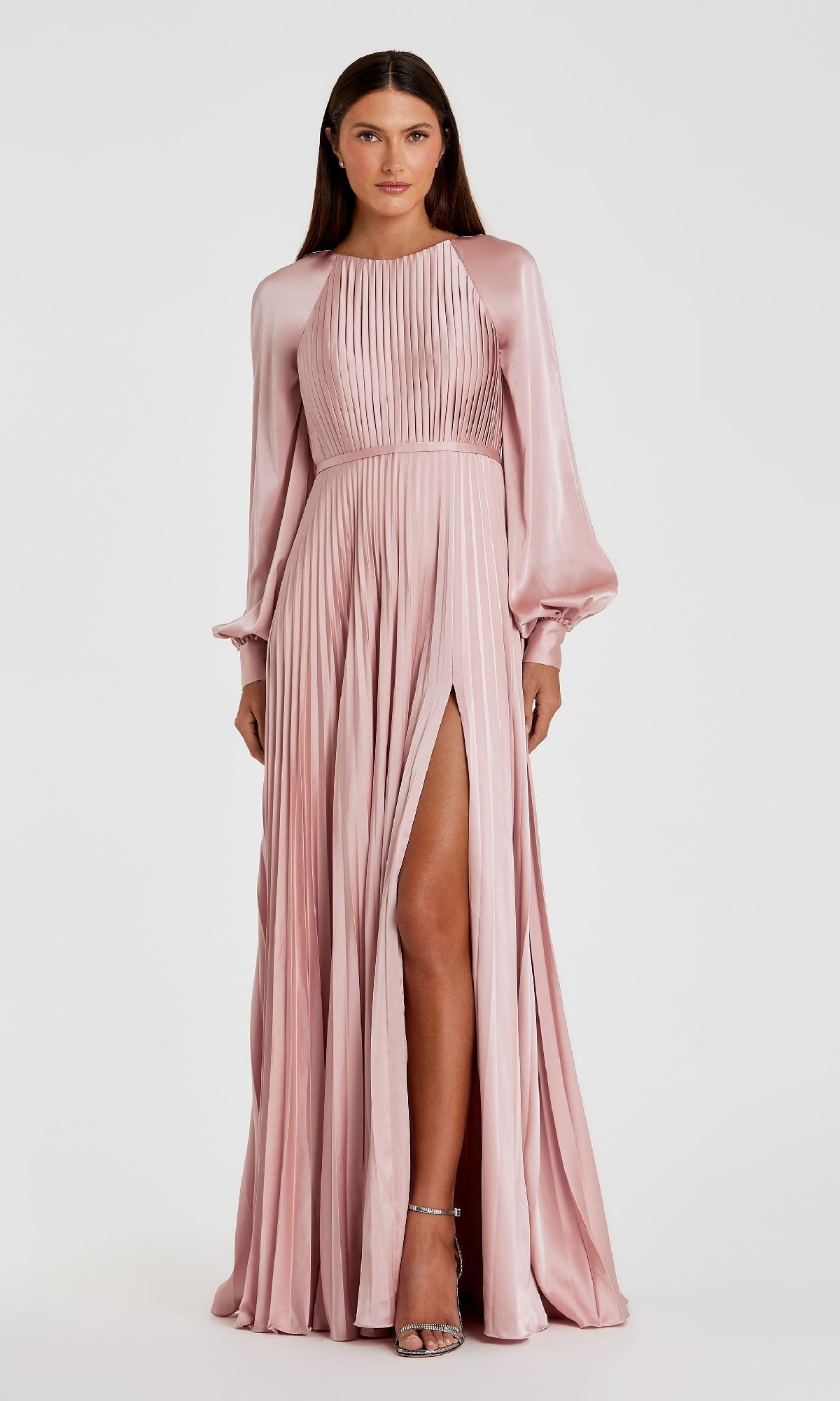 A woman models the Mac Duggal 26590, a light pink, long-sleeve pleated formal dress with a high slit and A-line silhouette, styled as an elegant evening gown against a plain white background.
