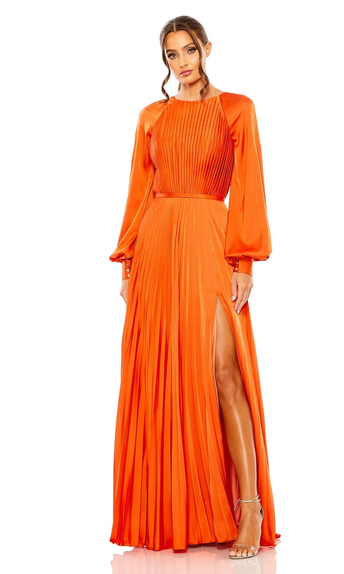 A woman stands gracefully against a white background wearing the Mac Duggal 26590: a vibrant orange long formal dress with long sleeves, pleated details, and a high slit, paired with strappy heels.
