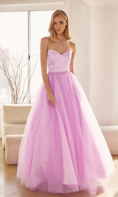 A woman stands indoors in the Strapless Sweetheart Long Prom Ball Gown 265, featuring a fitted bodice, corset-style back, and full light pink tulle skirt. Natural light streams through a window behind her.