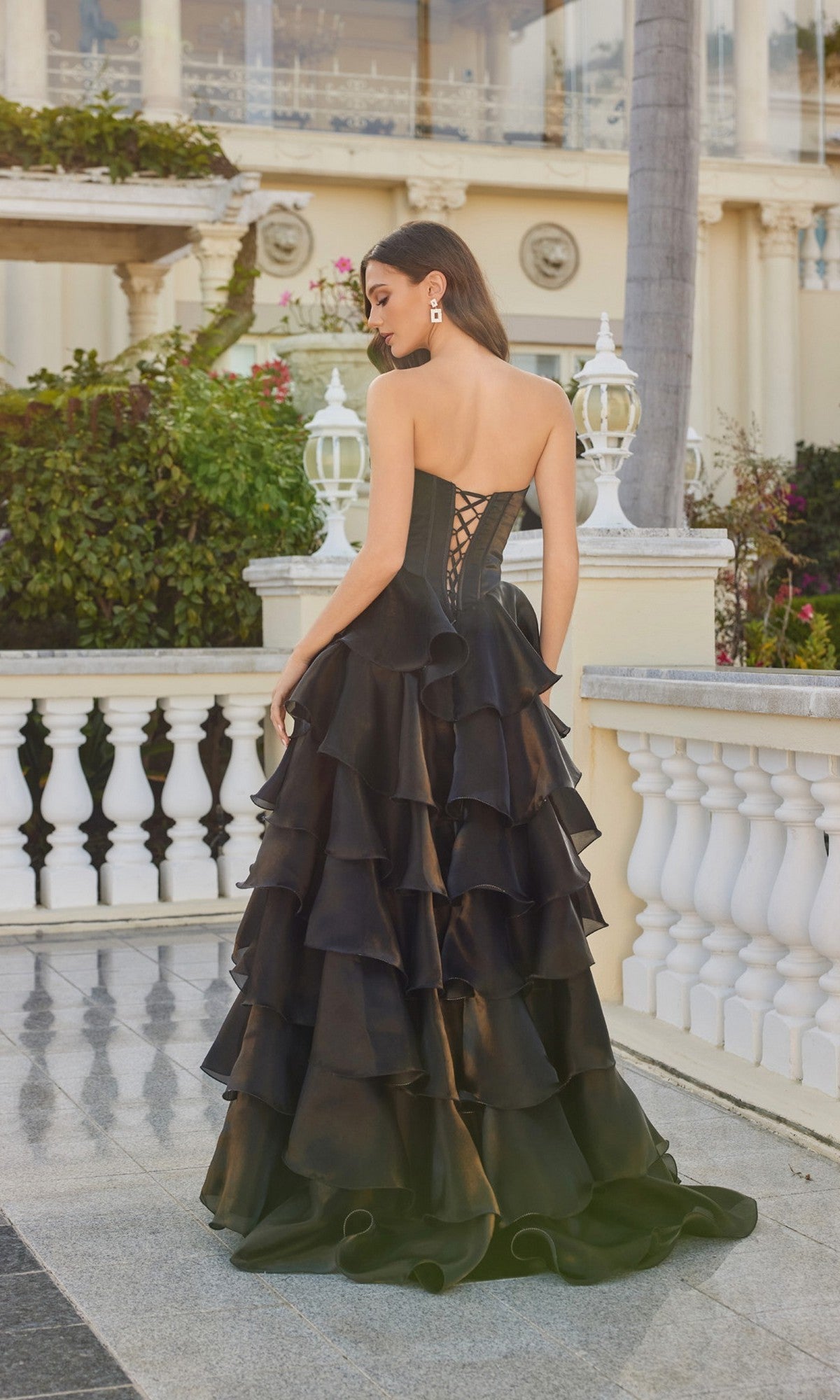 A woman wearing the Juliet JT2664S long prom dress, featuring a strapless corset bodice and tiered A-line skirt, stands outdoors near a white railing with her back to the camera.