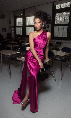 In a classroom, a person wears the Mac Duggal 26654 one-shoulder long formal dress in magenta, paired with silver heels and holding a small black handbag.