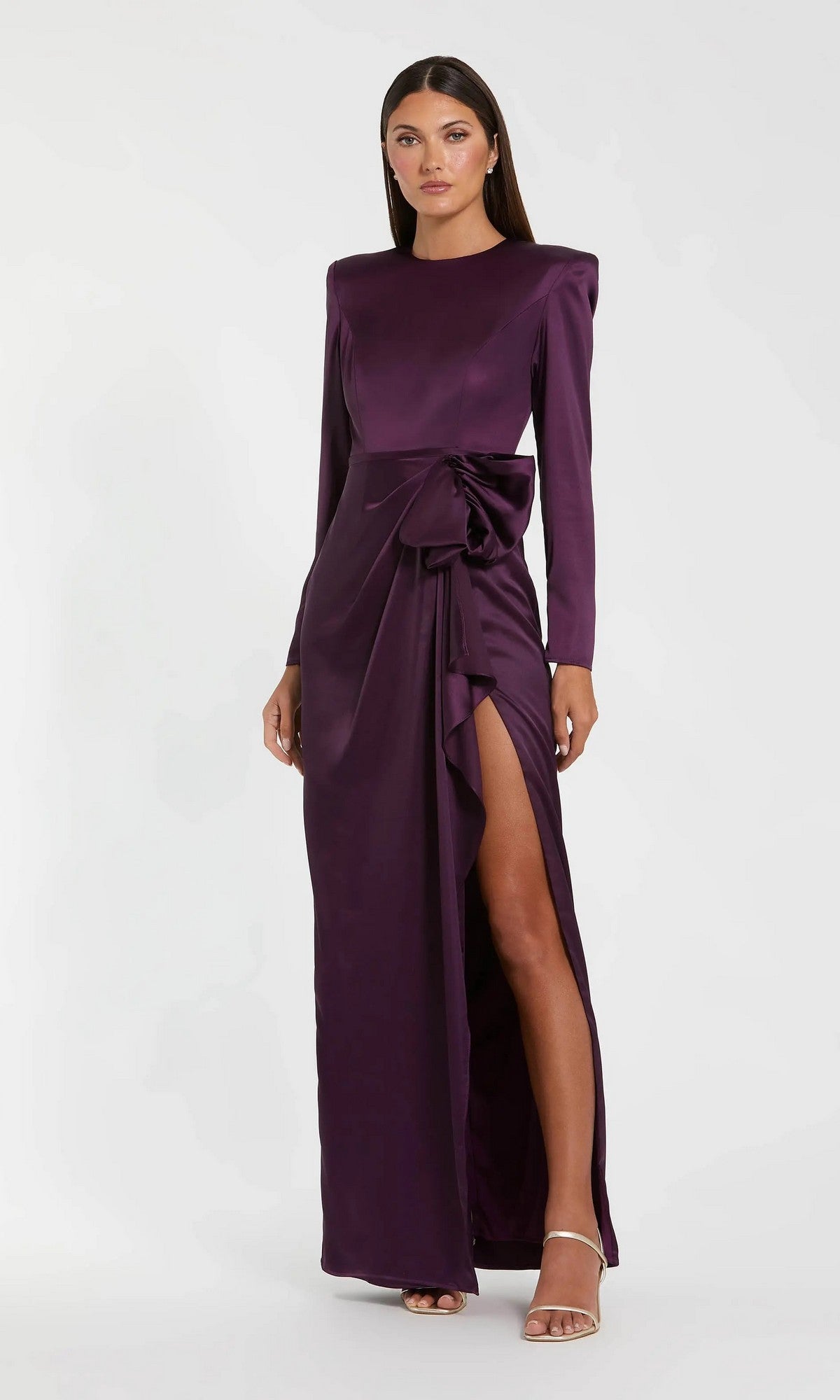 A woman in the Mac Duggal 26676 Bold-Shoulder Long Formal Dress—a bra-friendly deep purple gown with long sleeves, a side slit, and waist bow—stands against a plain light background.
