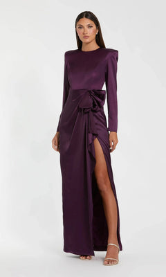 A woman in the Mac Duggal 26676 Bold-Shoulder Long Formal Dress—a bra-friendly deep purple gown with long sleeves, a side slit, and waist bow—stands against a plain light background.