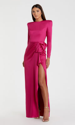 A woman models the Mac Duggal 26676 Bold-Shoulder Long Formal Dress, a vibrant pink, bra-friendly evening gown with long sleeves, a side slit, and a waist bow, posed against a simple white background.