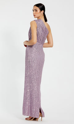 A woman stands in a studio wearing the Mac Duggal 26694 one-shoulder floor-length sequin formal dress in lavender, paired with beige heels, looking over her shoulder.