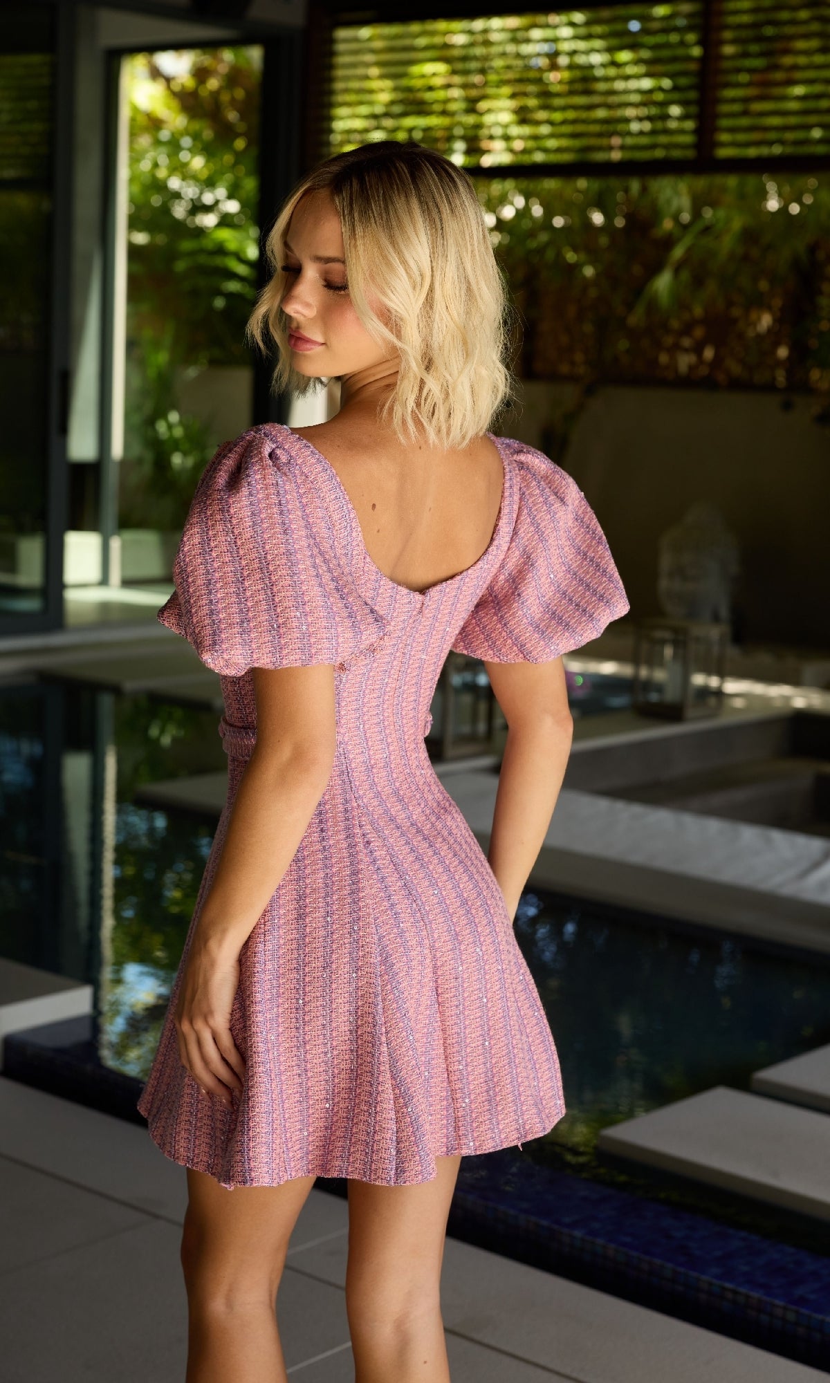 A blonde woman stands by an indoor pool, wearing the Ava Presley 26704 short-sleeve short homecoming dress in textured light purple and looking over her shoulder.