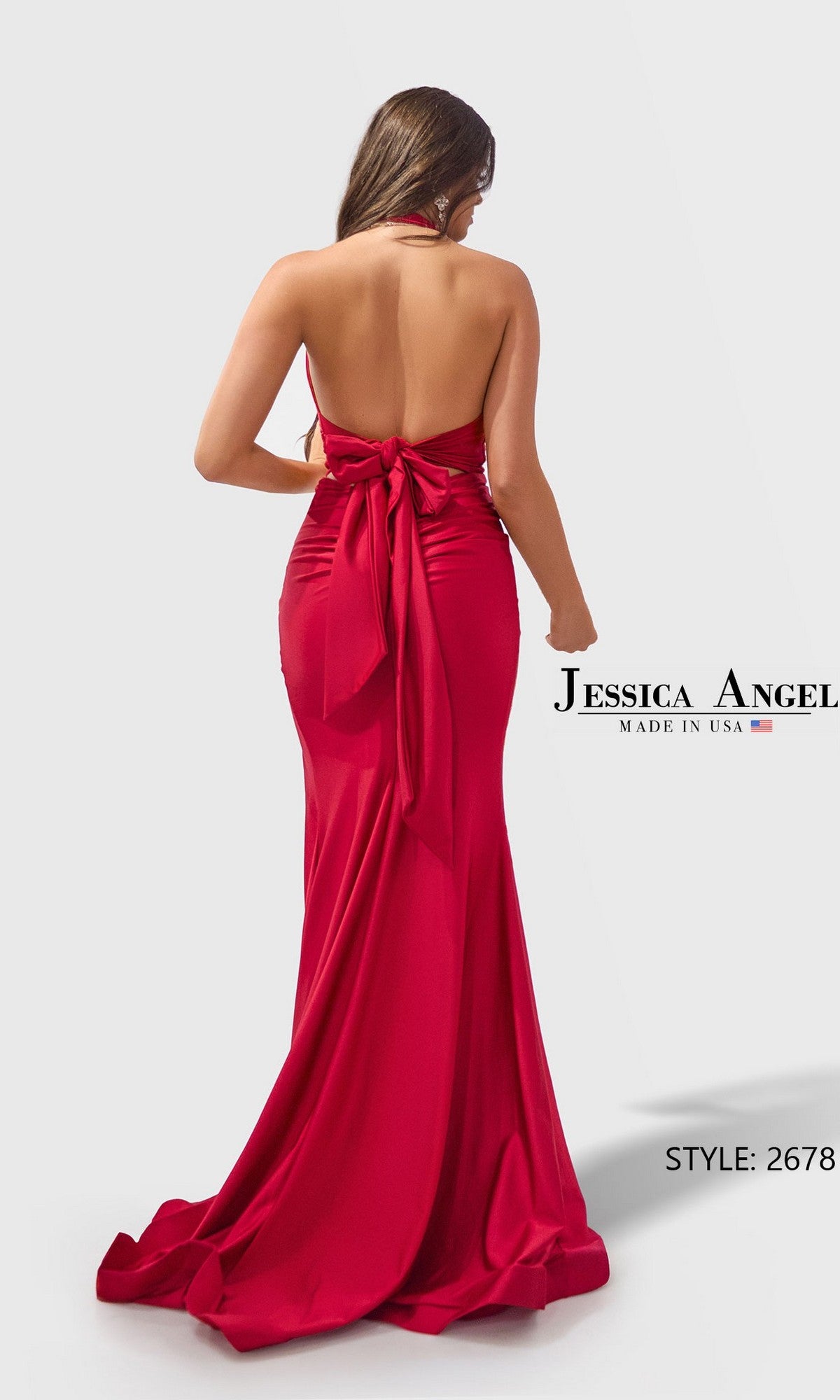 A woman stands against a plain light background wearing the Jessica Angel 2678, a long red jersey prom dress featuring a fitted sheath silhouette, floor-length hem, backless design, and a large bow accent at the lower back.