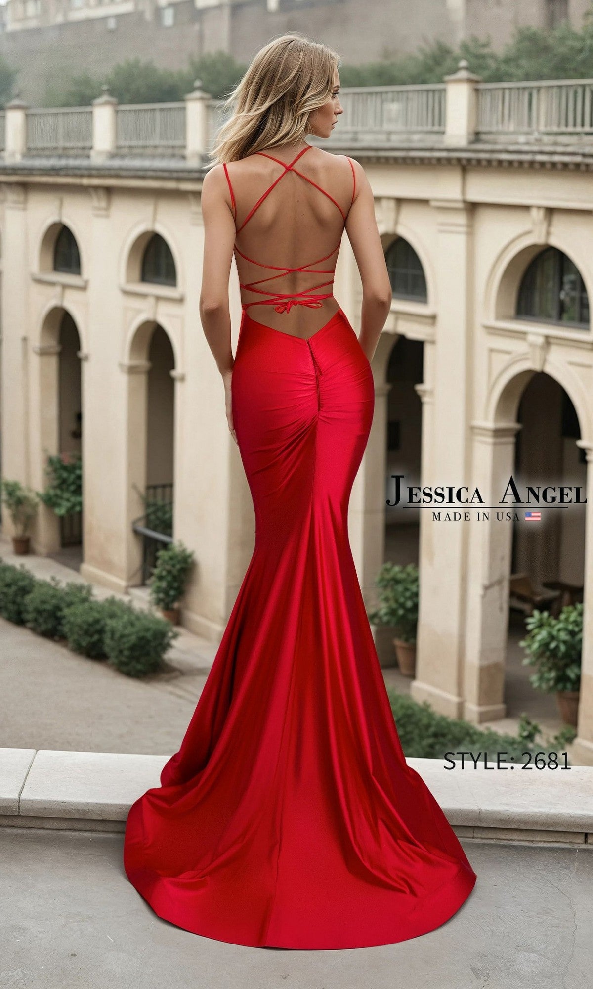 A woman in a form-fitting, satin red mermaid prom dress with crisscross straps and an open back stands on a terrace. She wears the Long Prom Dress: Jessica Angel 2681.
