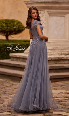 Woman in a floor-length, short-sleeve lavender La Femme 26893 gown with a lace embroidered bodice stands outdoors on stone pavement near a white stone structure, showcasing this elegant long formal dress—an ideal choice as a mother of the bride dress.