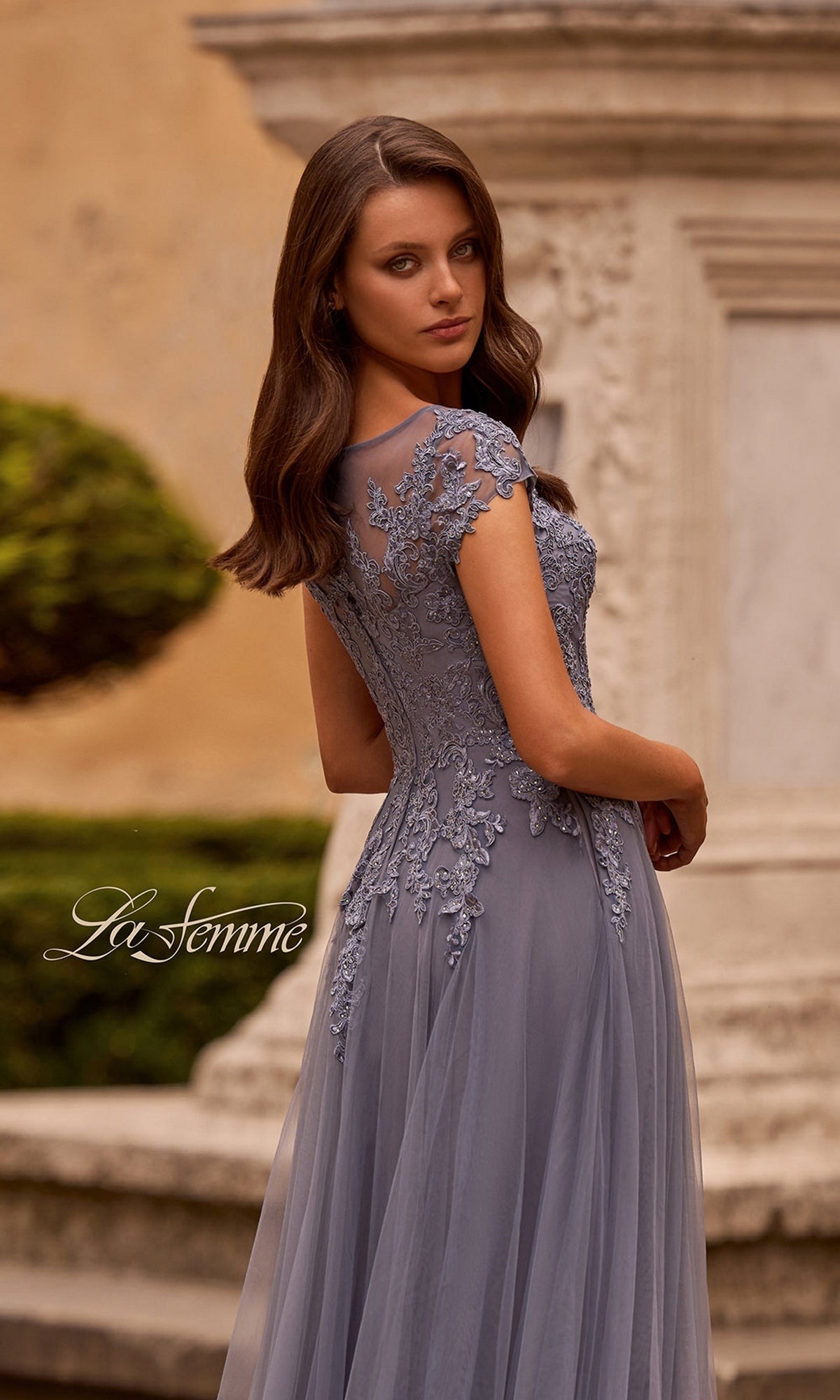 A woman stands outdoors near a stone structure, looking over her shoulder in the La Femme 26893 long formal dress—a short-sleeved lavender lace gown featuring a beautifully embroidered lace bodice. The image displays the text “La Femme.”.