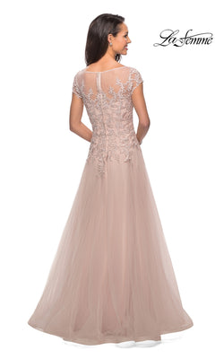 A woman is seen standing and facing away from the camera, wearing the La Femme 26893 long formal dress— a floor-length, short-sleeved gown in blush pink, featuring a lace embroidered bodice and delicate appliqué details on the upper back.